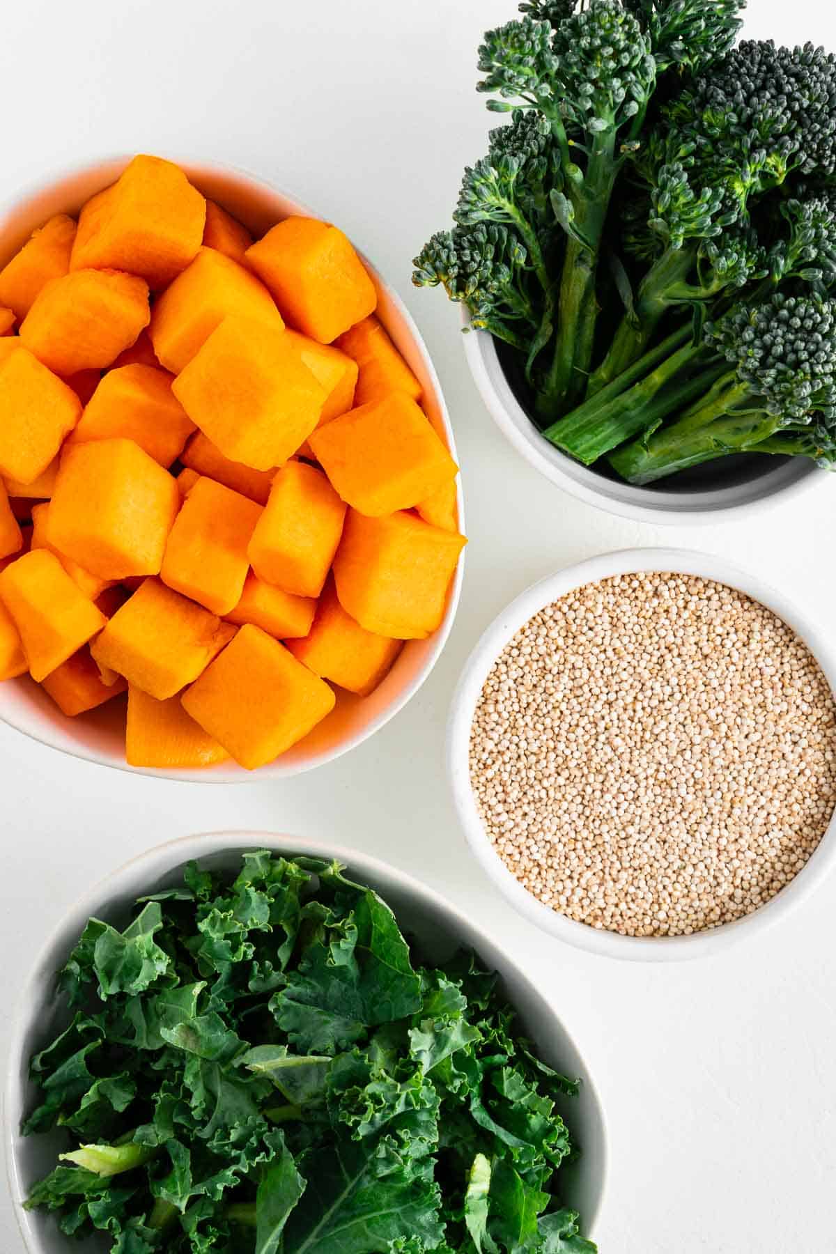 four white bowls filled with cubed butternut squash, broccolini, quinoa, and chopped kale