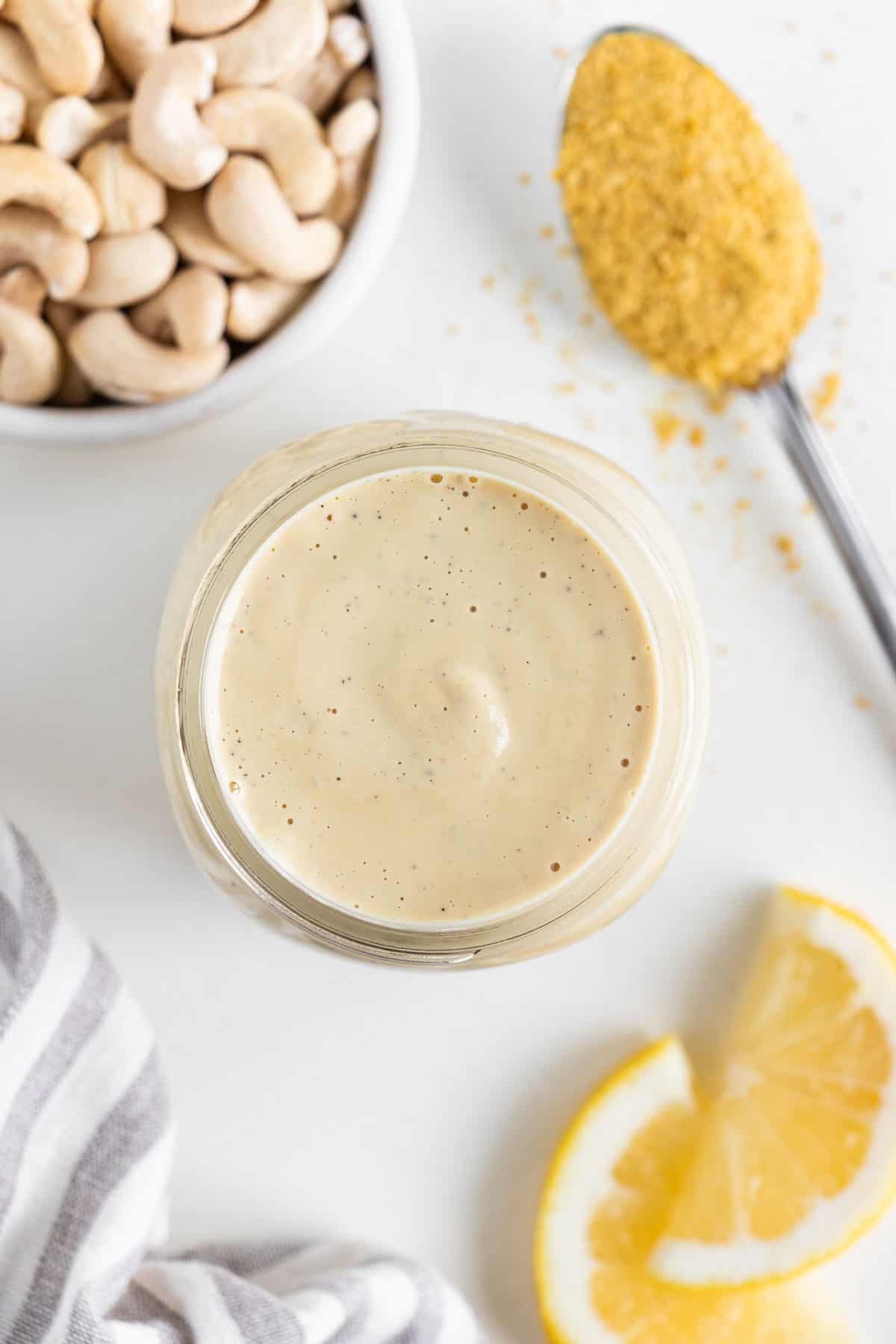 an overhead photo of vegan cashew cream sauce inside a mason jar, surrounded by a bowl of cashews, lemon slices, and a spoonful of nutritional yeast