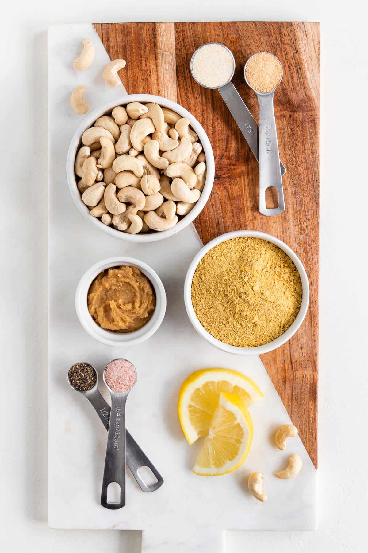 a wood and marble cutting board topped with bowls of cashews, nutritional yeast, white miso paste, salt, pepper, and lemon slices