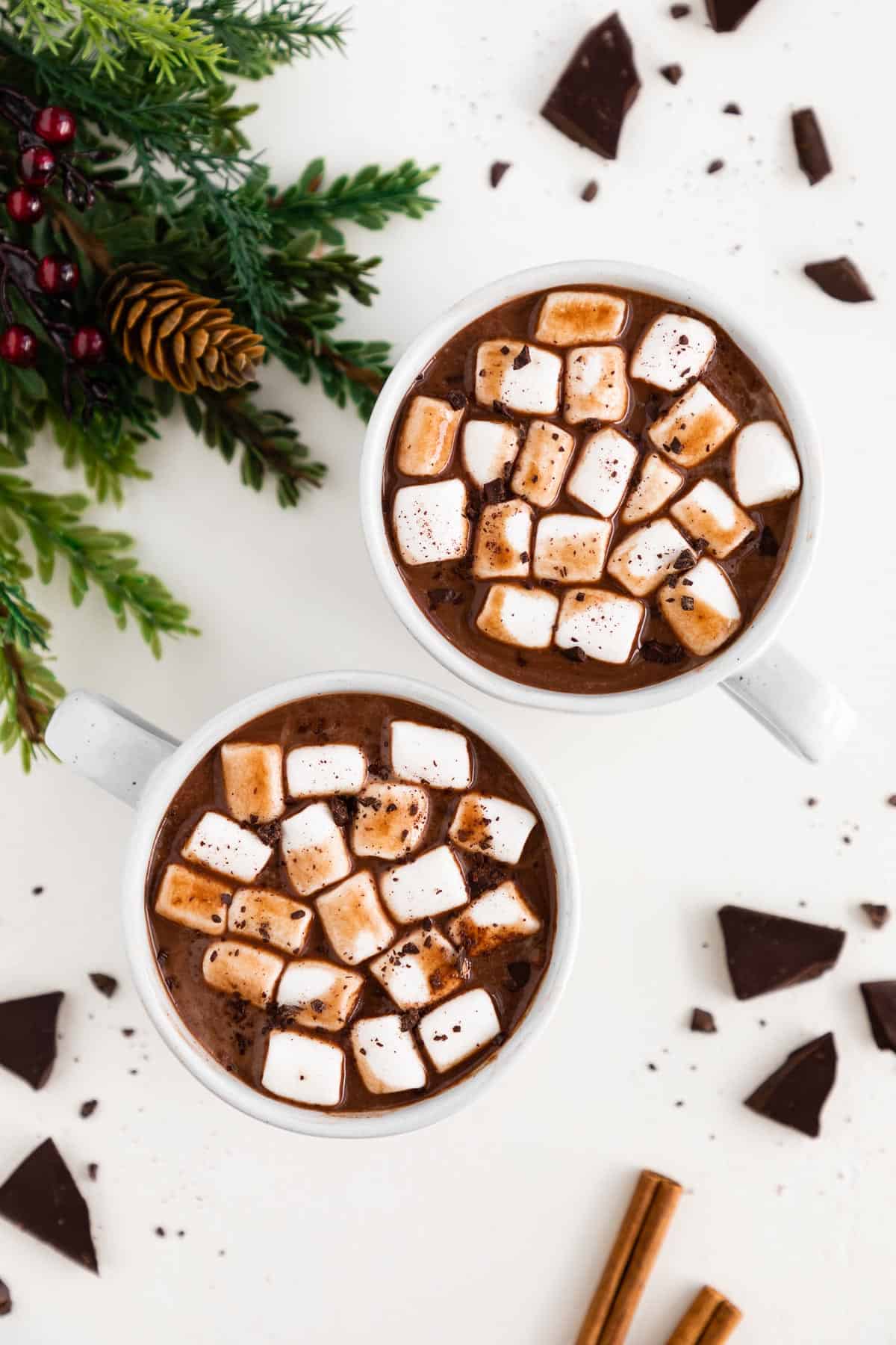 two white mugs filled with vegan hot chocolate, mini marshmallows, and chocolate shavings, surrounded by pieces of dark chocolate