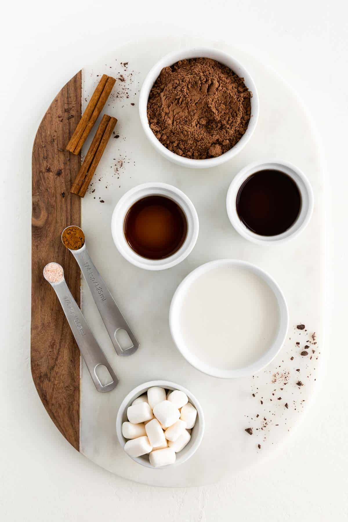 a marble board topped with bowls of cocoa powder, oat milk, vanilla extract, maple syrup, marshmallows, and cinnamon sticks