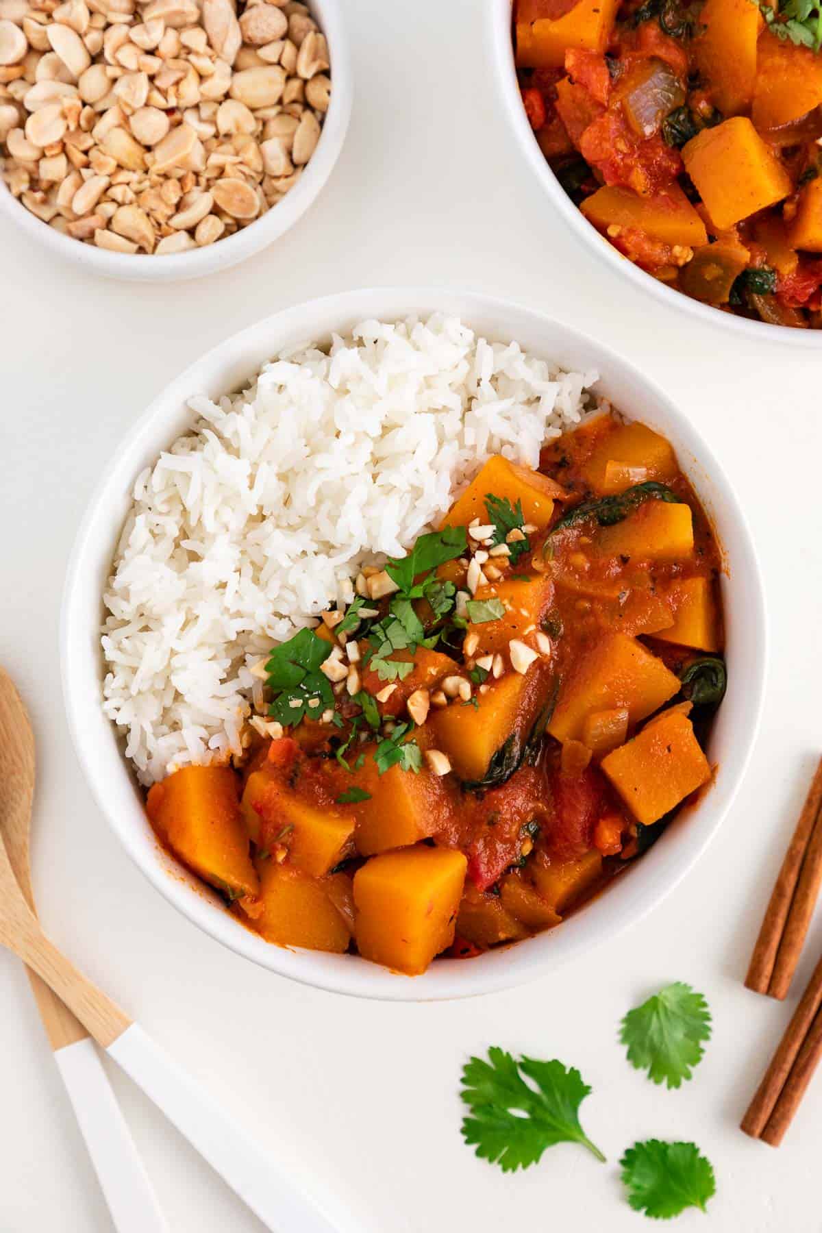 a bowl of butternut squash curry and white rice surrounded by another bowl of curry, a bowl filled with crushed peanuts, wooden spoons, and cinnamon sticks