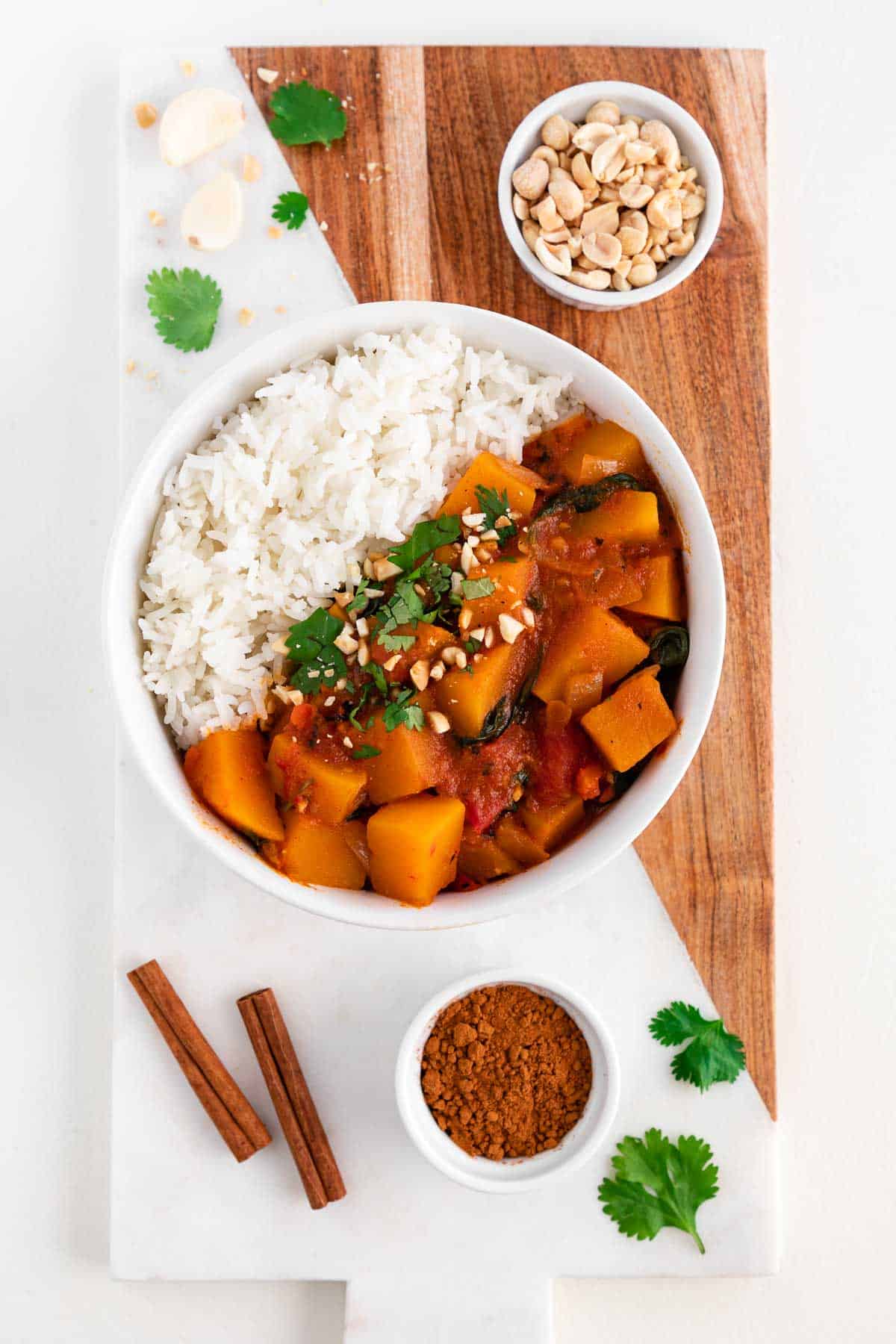 a wood and marble cutting board topped with a bowl of butternut squash curry, a bowl of peanuts, a bowl of cinnamon, and fresh cilantro