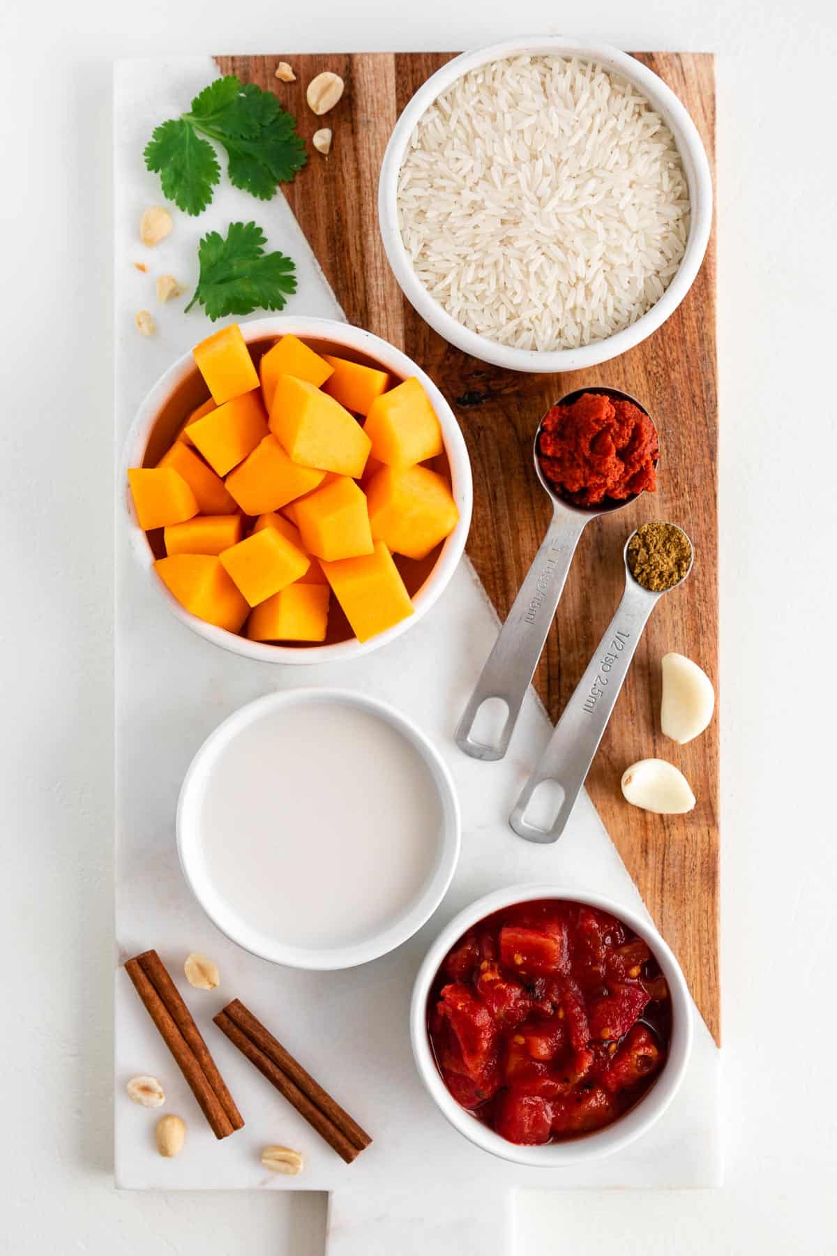 a marble and wood cutting board topped with bowls of coconut milk, diced tomatoes, squash, white rice, cinnamon sticks, red curry paste, cumin, and cilantro