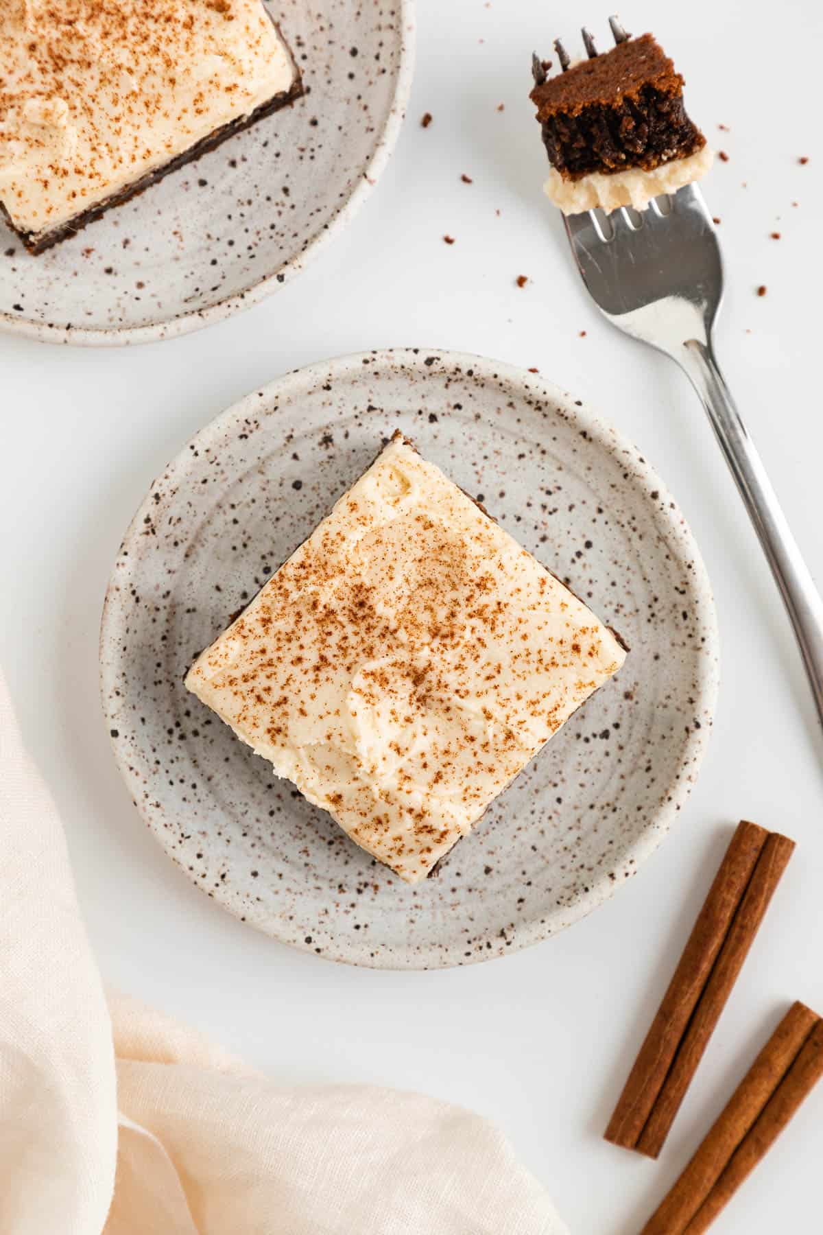 a vegan gingerbread cookie bar with frosting on a ceramic plate, beside a fork and cinnamon sticks