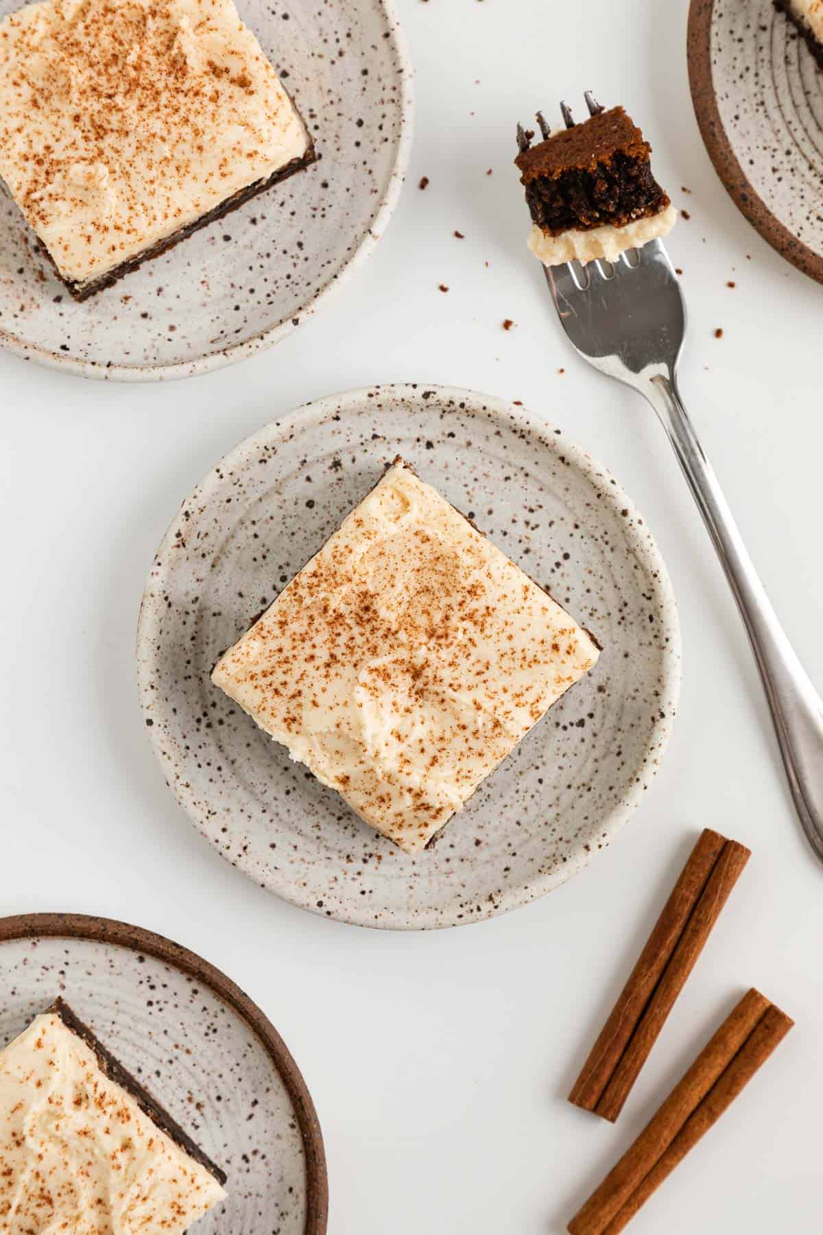 three vegan frosted gingerbread cookie bars on ceramic dessert plates, beside a fork and two cinnamon sticks