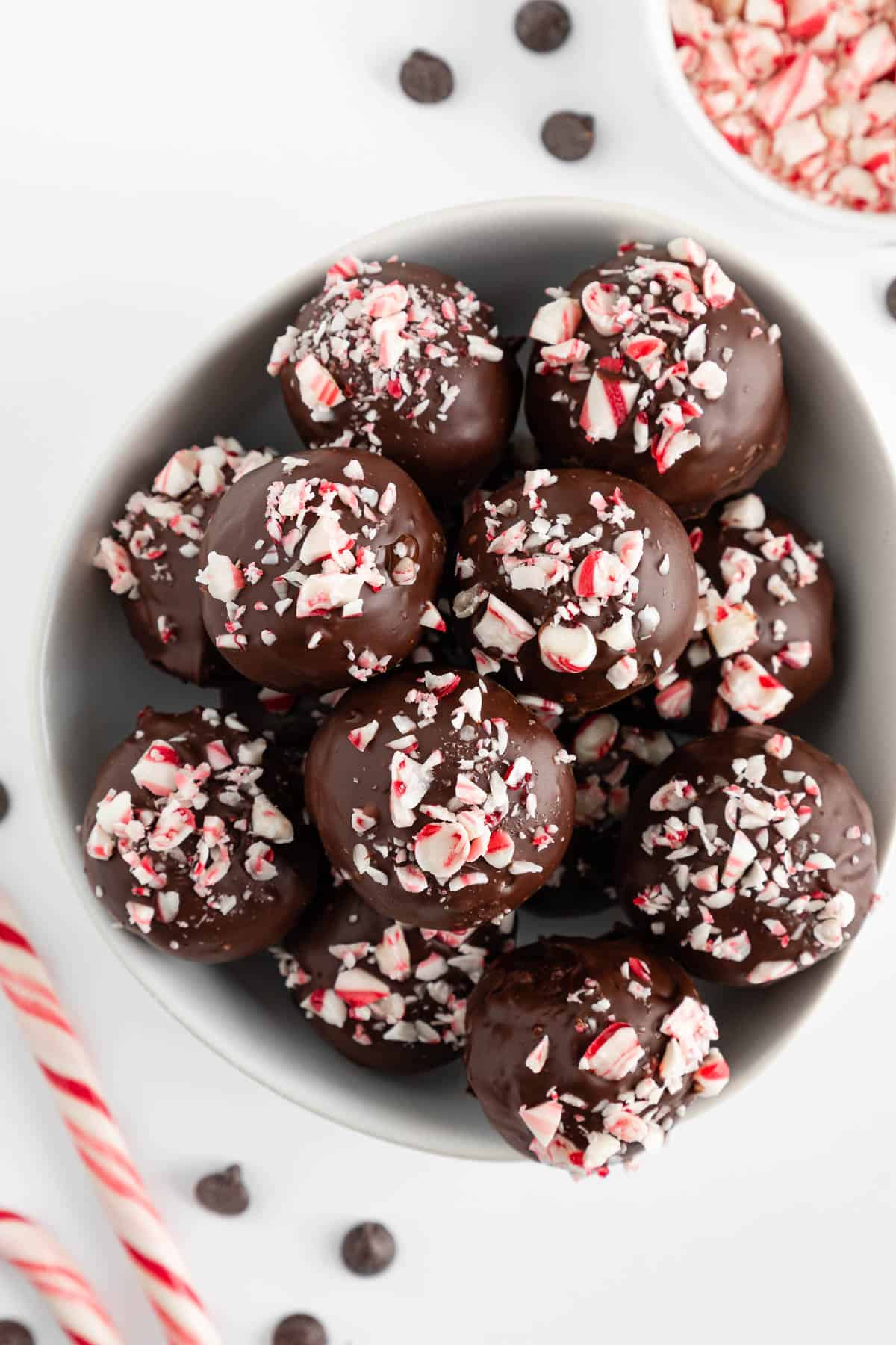 healthy chocolate peppermint truffles stacked inside a white bowl, beside a bowl of crushed candy canes and chocolate chips