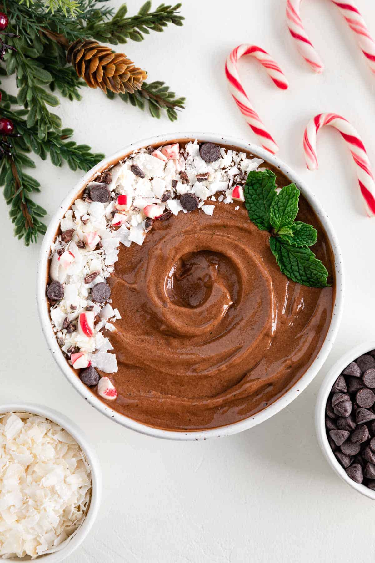 peppermint mocha smoothie bowl surrounded by candy canes, a bowl of chocolate chips, a bowl of coconut flakes, and a holiday leaf
