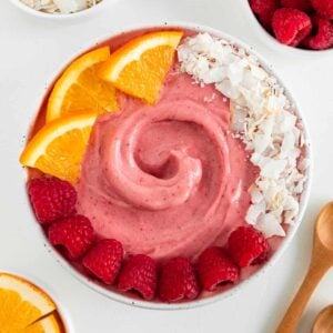 a raspberry orange smoothie bowl surrounded by small white bowls filled with coconut flakes, berries, orange slices, and two wooden spoons