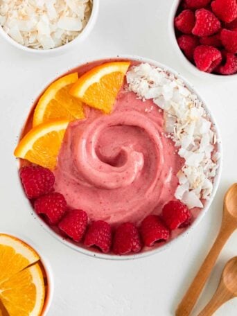 a raspberry orange smoothie bowl surrounded by small white bowls filled with coconut flakes, berries, orange slices, and two wooden spoons
