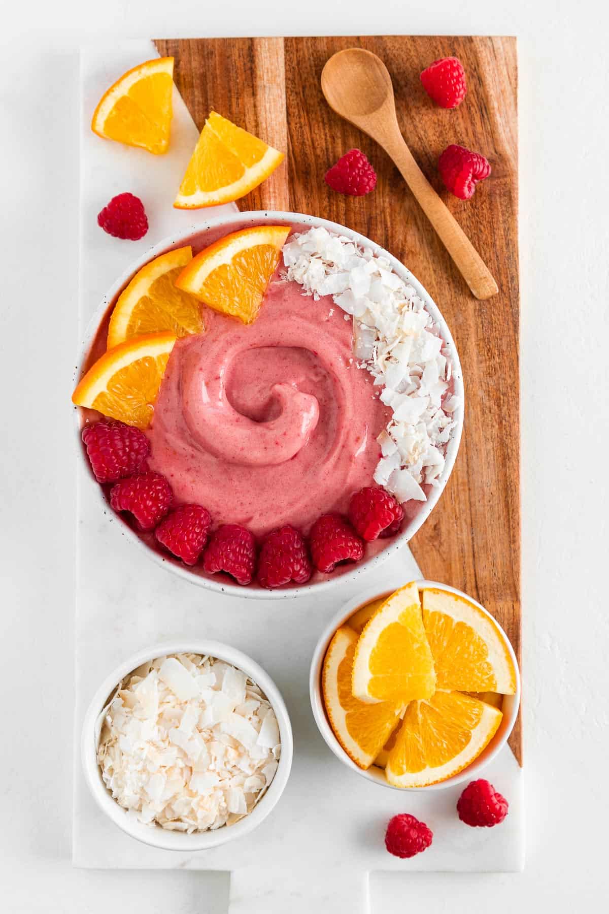 a wood and marble board topped with a raspberry orange smoothie bowl, wooden spoon, and bowls filled with fresh fruit