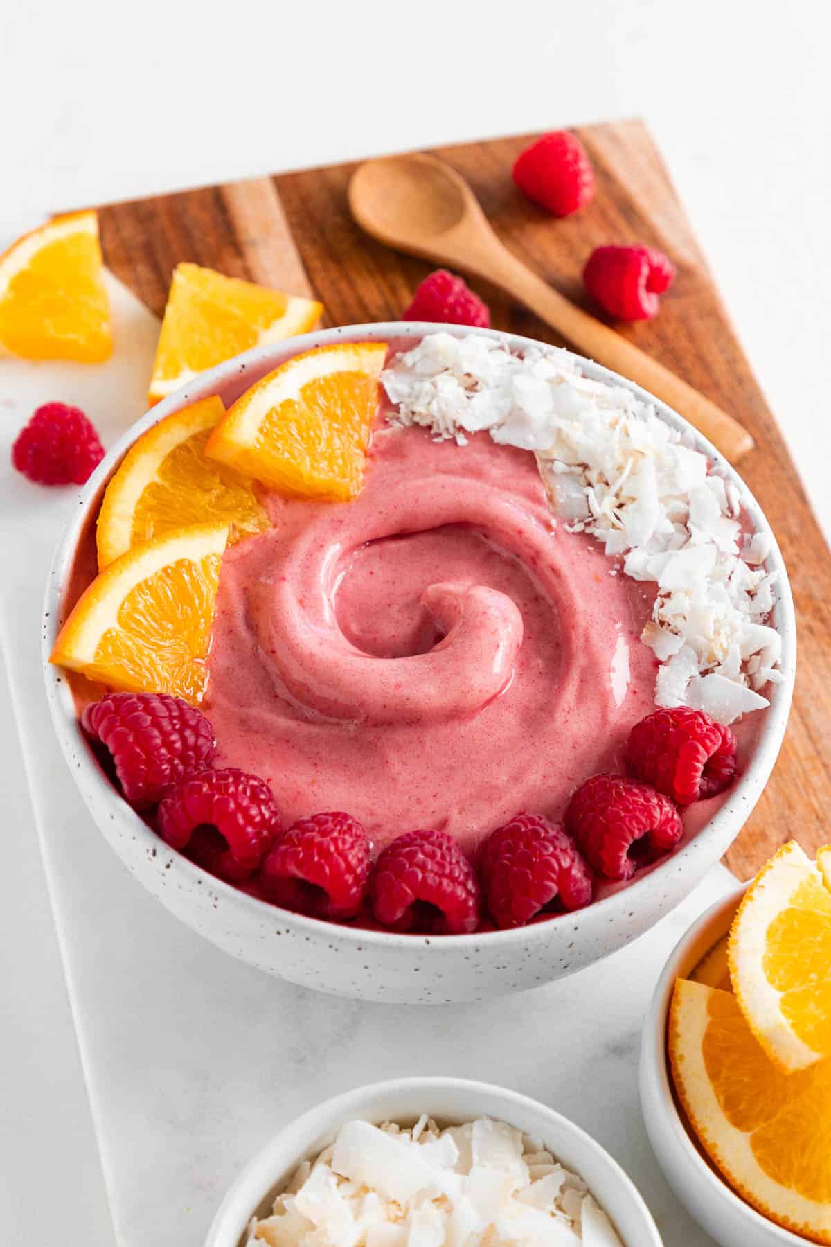 a raspberry smoothie inside a ceramic bowl, surrounded by orange slices, coconut flakes, and berries