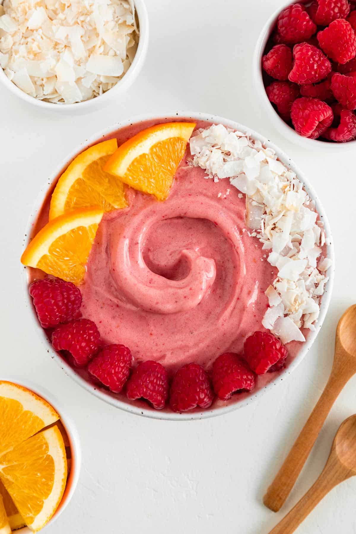 a raspberry orange smoothie bowl surrounded by small white bowls filled with coconut flakes, berries, orange slices, and two wooden spoons