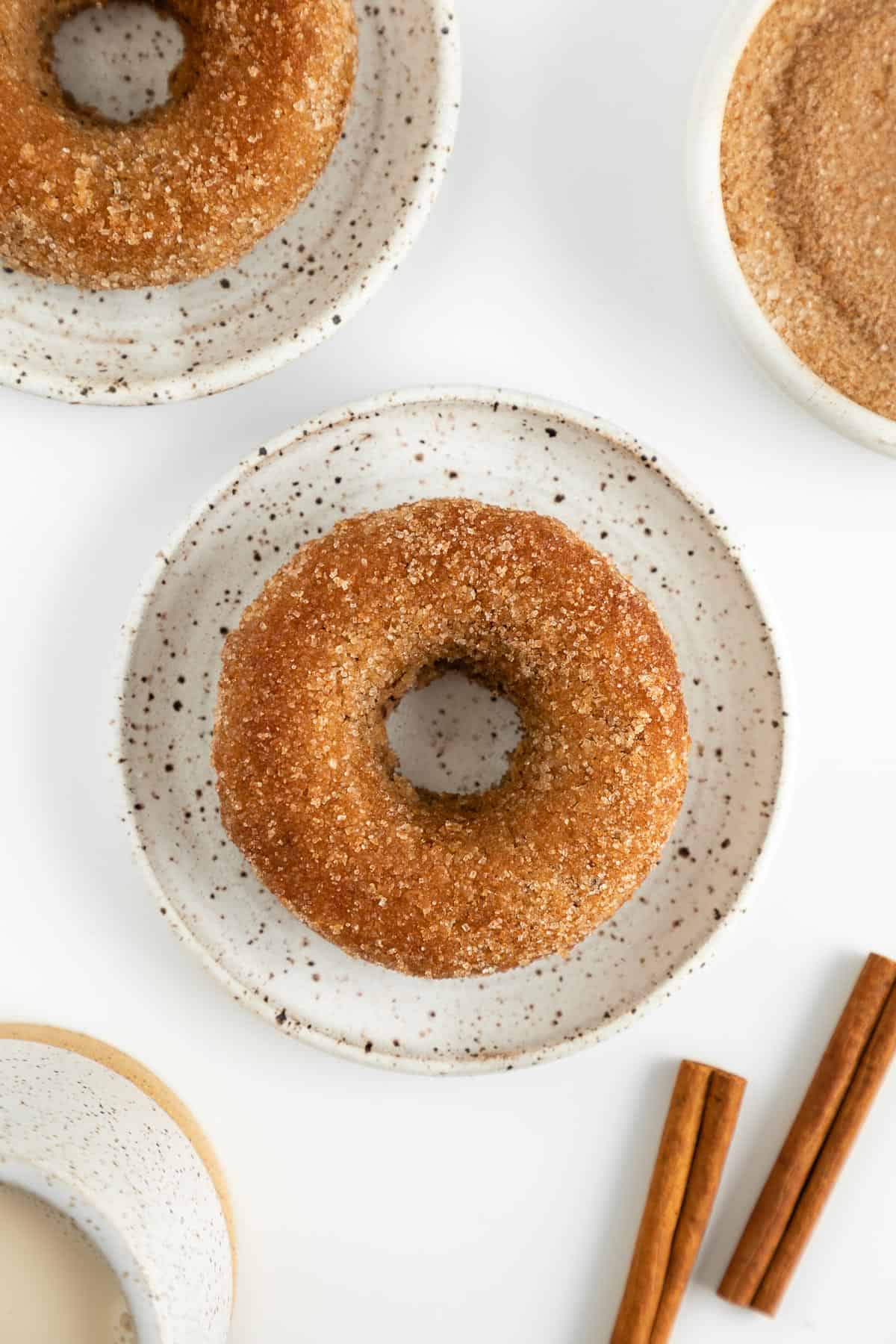 a vegan cinnamon sugar donut on a small ceramic plate beside a glass of almond milk, cinnamon sticks, and a bowl of cinnamon