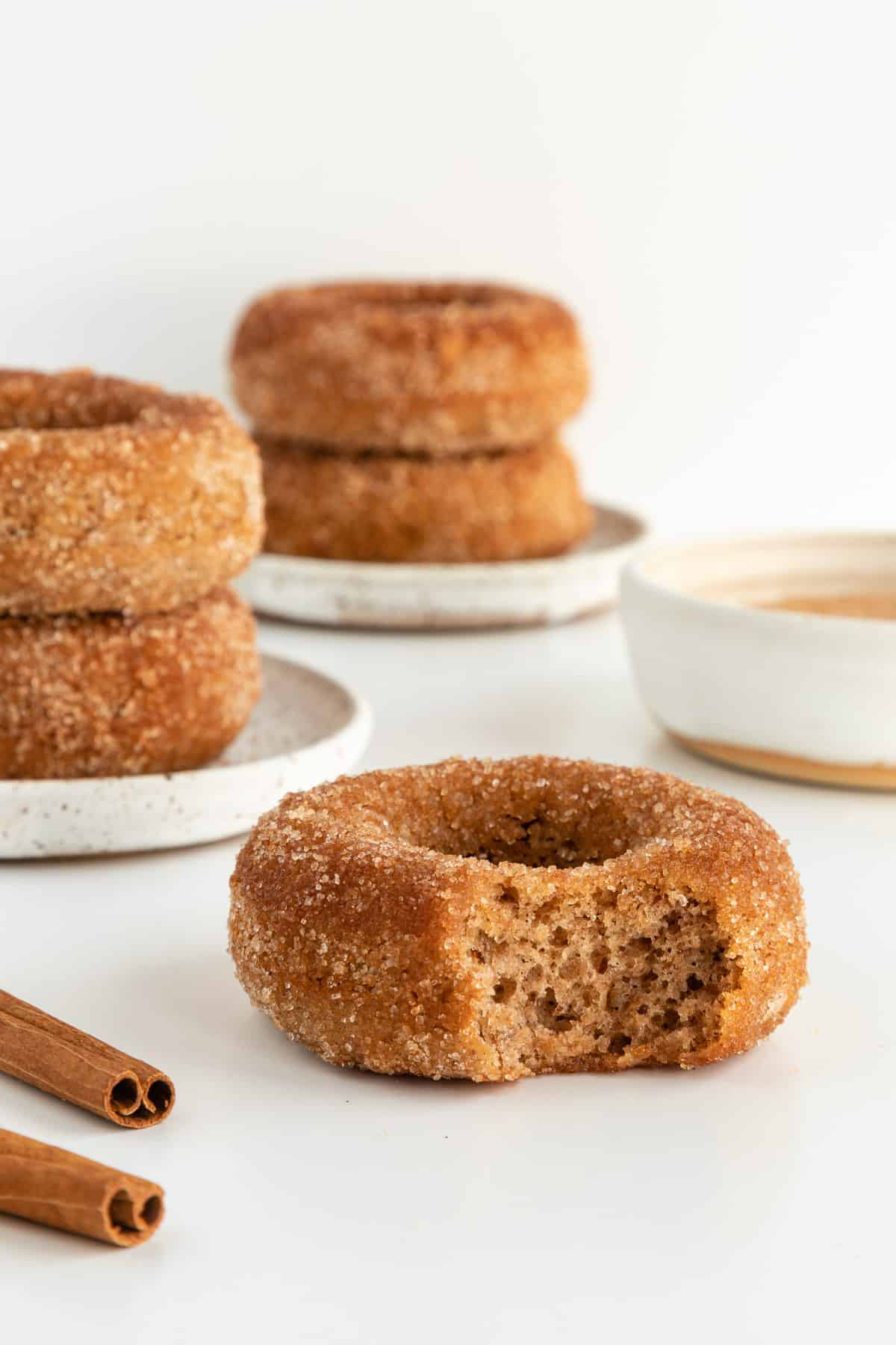 a partially bitten vegan cinnamon sugar donut with stacks of donuts behind it