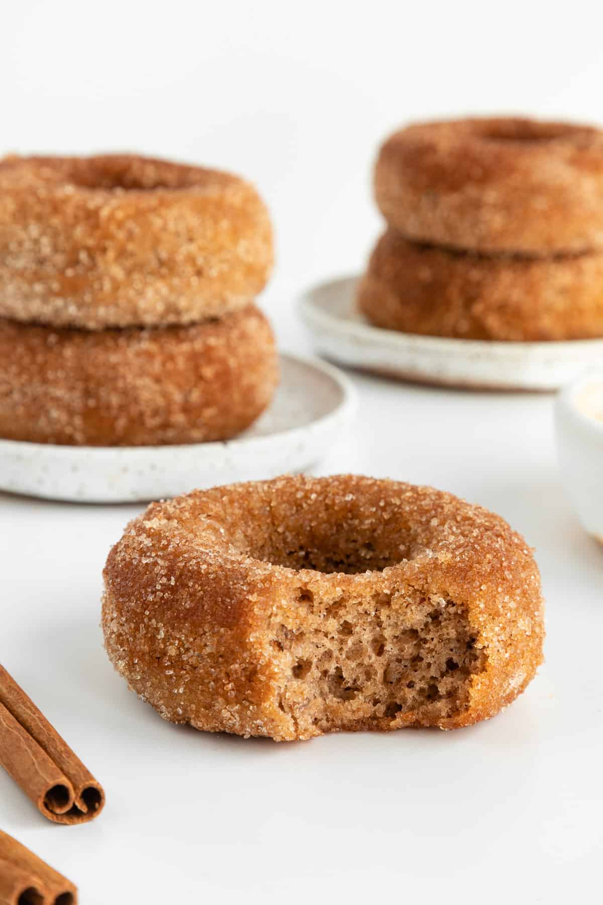 a partially bitten vegan cinnamon sugar donut with stacks of donuts behind it