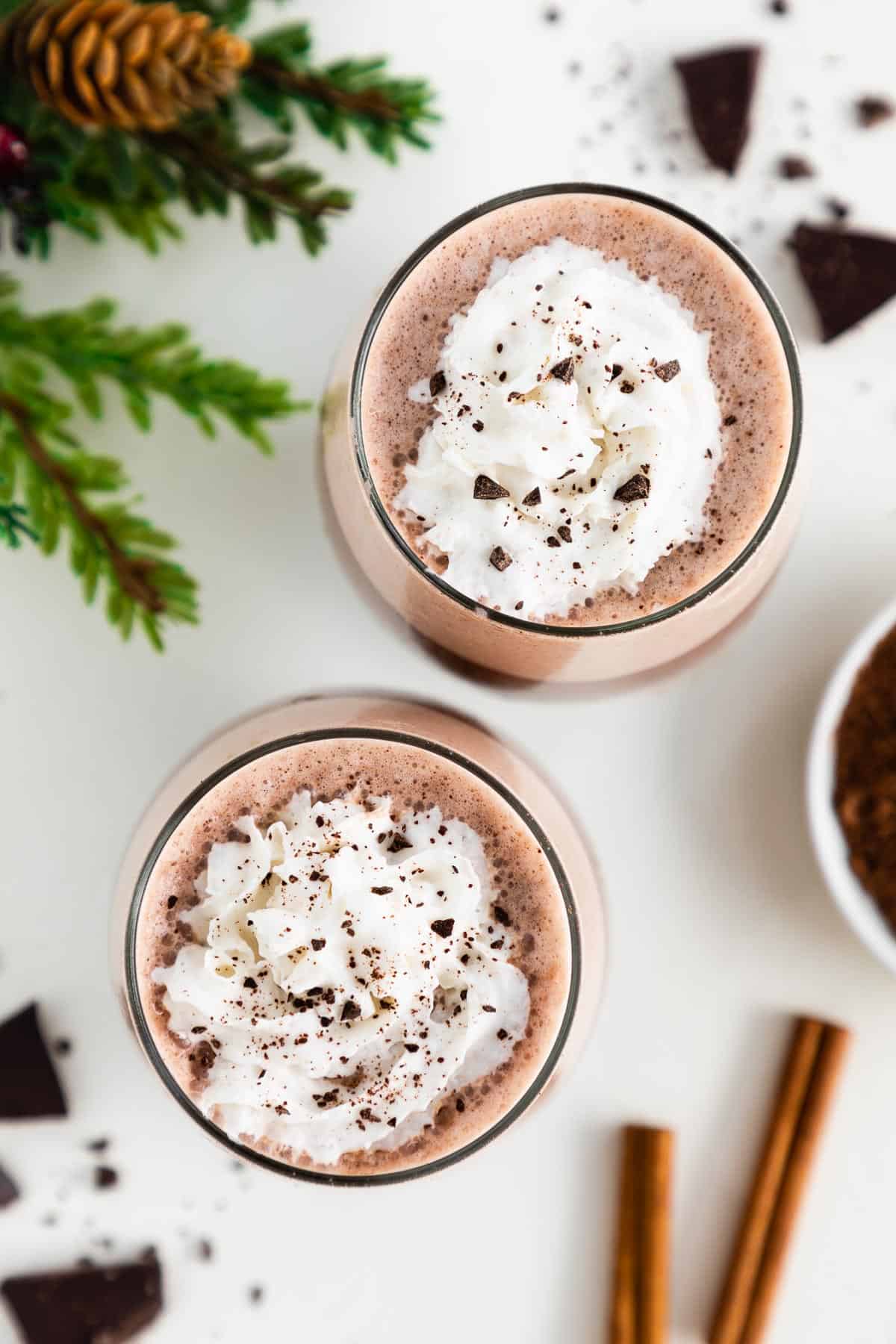 vegan hot chocolate in two glasses with whipped cream on top, surrounded by pieces of dark chocolate, cinnamon sticks, and a bowl of cocoa powder