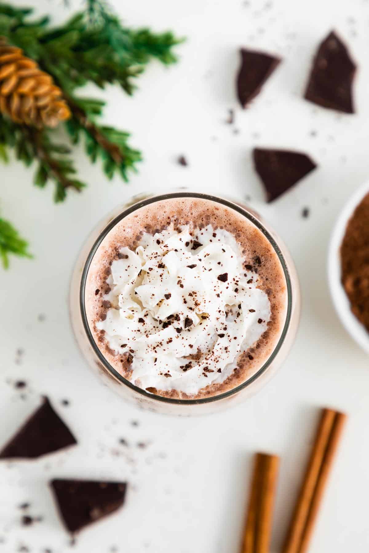 an overhead photo of vegan frozen hot chocolate in a glass with whipped cream, surrounded by pieces of dark chocolate, cinnamon sticks, and a bowl of cocoa powder