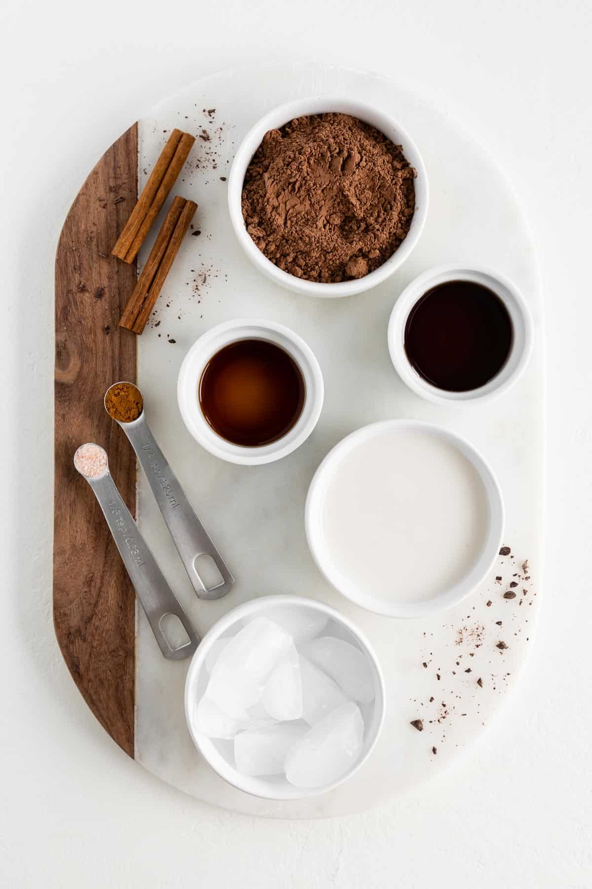 a marble and wood cutting board topped with bowls of cocoa powder, vanilla extract, maple syrup, almond milk, ice cubes, and cinnamon