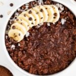 a white bowl filled with chocolate oatmeal and topped with banana, chocolate chips, coconut flakes, and cacao nibs