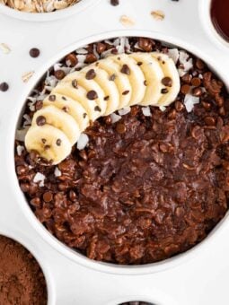 a white bowl filled with chocolate oatmeal and topped with banana, chocolate chips, coconut flakes, and cacao nibs