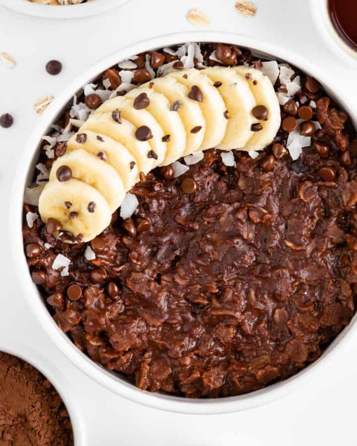a white bowl filled with chocolate oatmeal and topped with banana, chocolate chips, coconut flakes, and cacao nibs