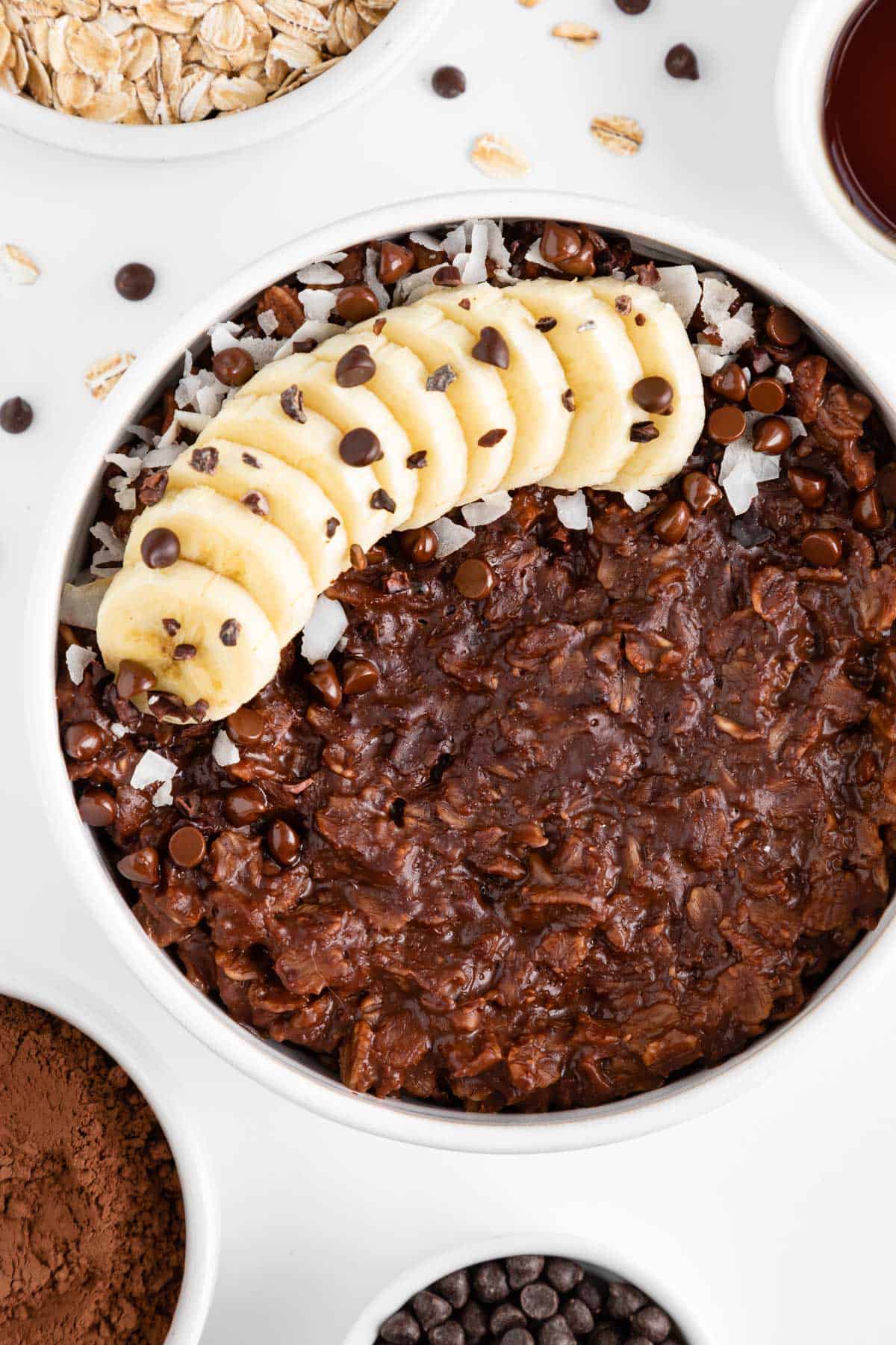 a white bowl filled with chocolate oatmeal and topped with banana, chocolate chips, coconut flakes, and cacao nibs