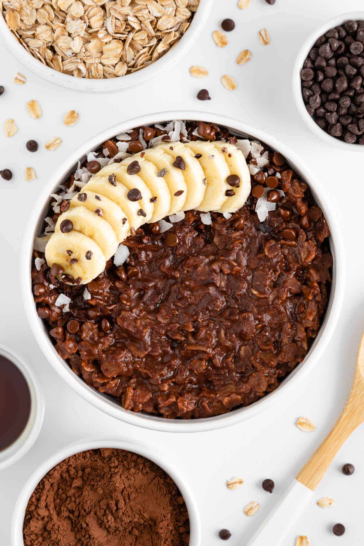 a bowl of chocolate oatmeal surrounded by small white bowls filled with cocoa powder, maple syrup, chocolate chips, and rolled oats