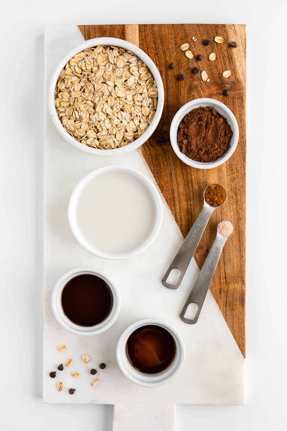 a marble and wood cutting board topped with bowls of rolled oats, cocoa powder, almond milk, maple syrup, vanilla extract, cinnamon, and salt