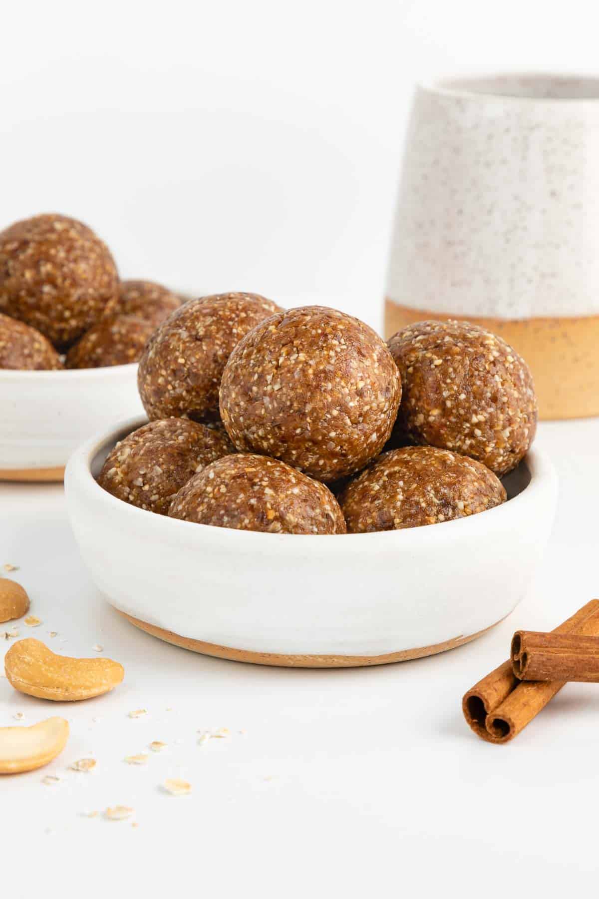 cinnamon roll energy balls stacked inside a small white bowl surrounded by cashews, oats, and cinnamon sticks