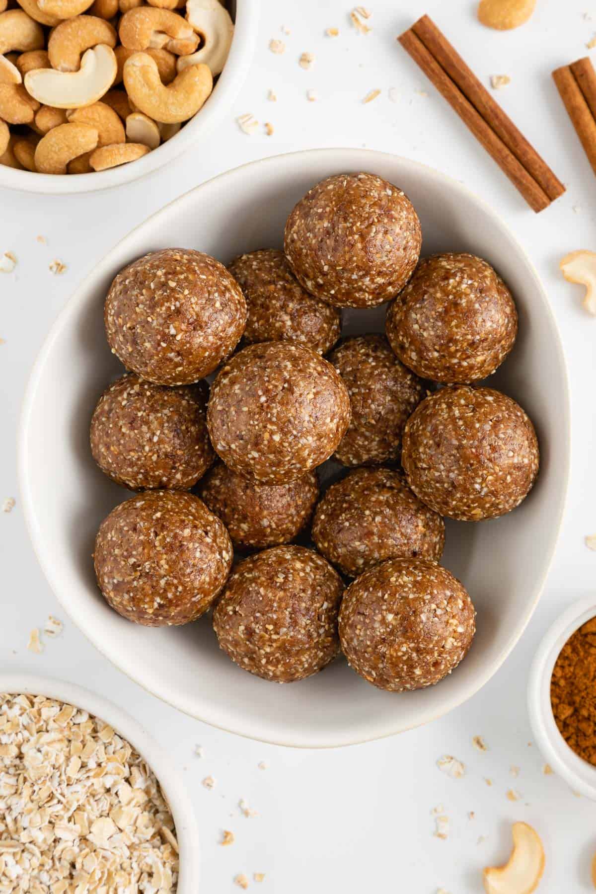 cinnamon roll energy balls inside a white bowl surrounded by oats, cashews, and cinnamon