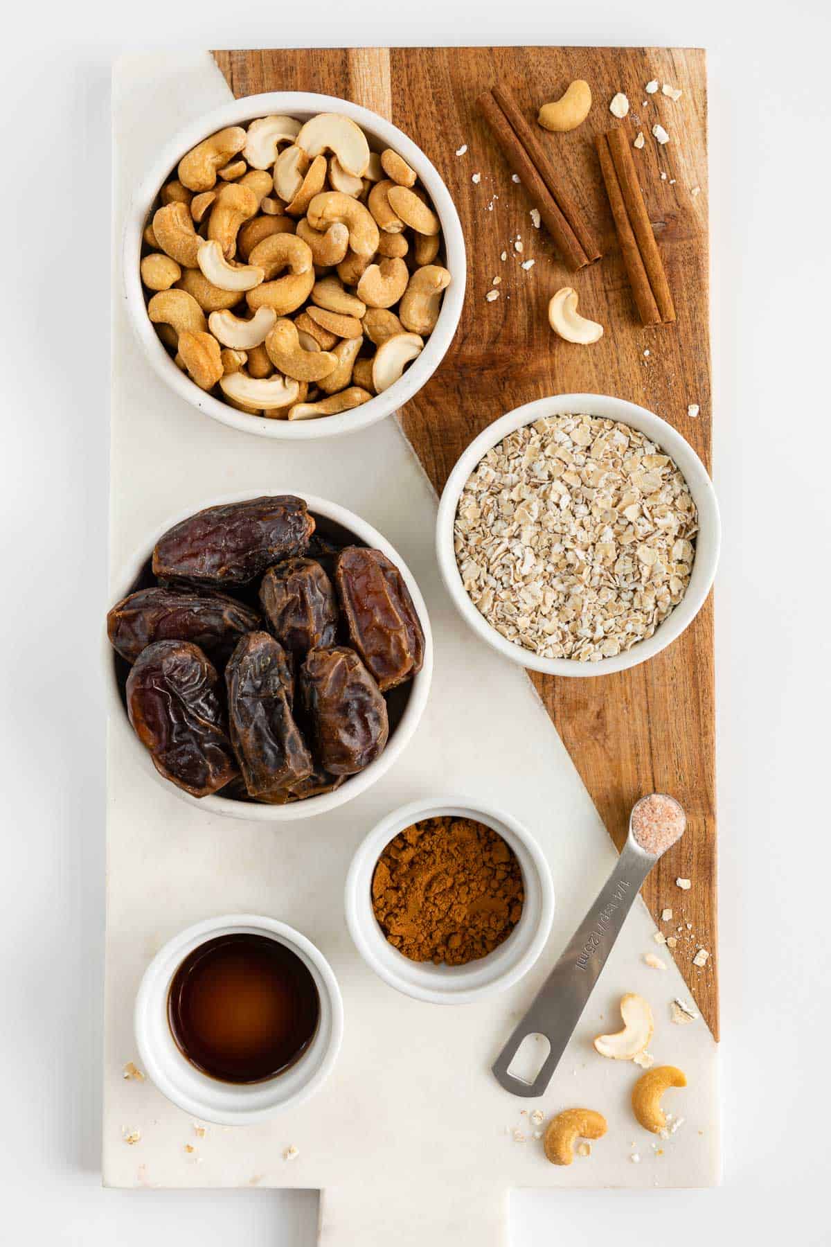 bowls filled with cashews, medjool dates, oats, cinnamon, vanilla extract, and salt on a marble and wood cutting board