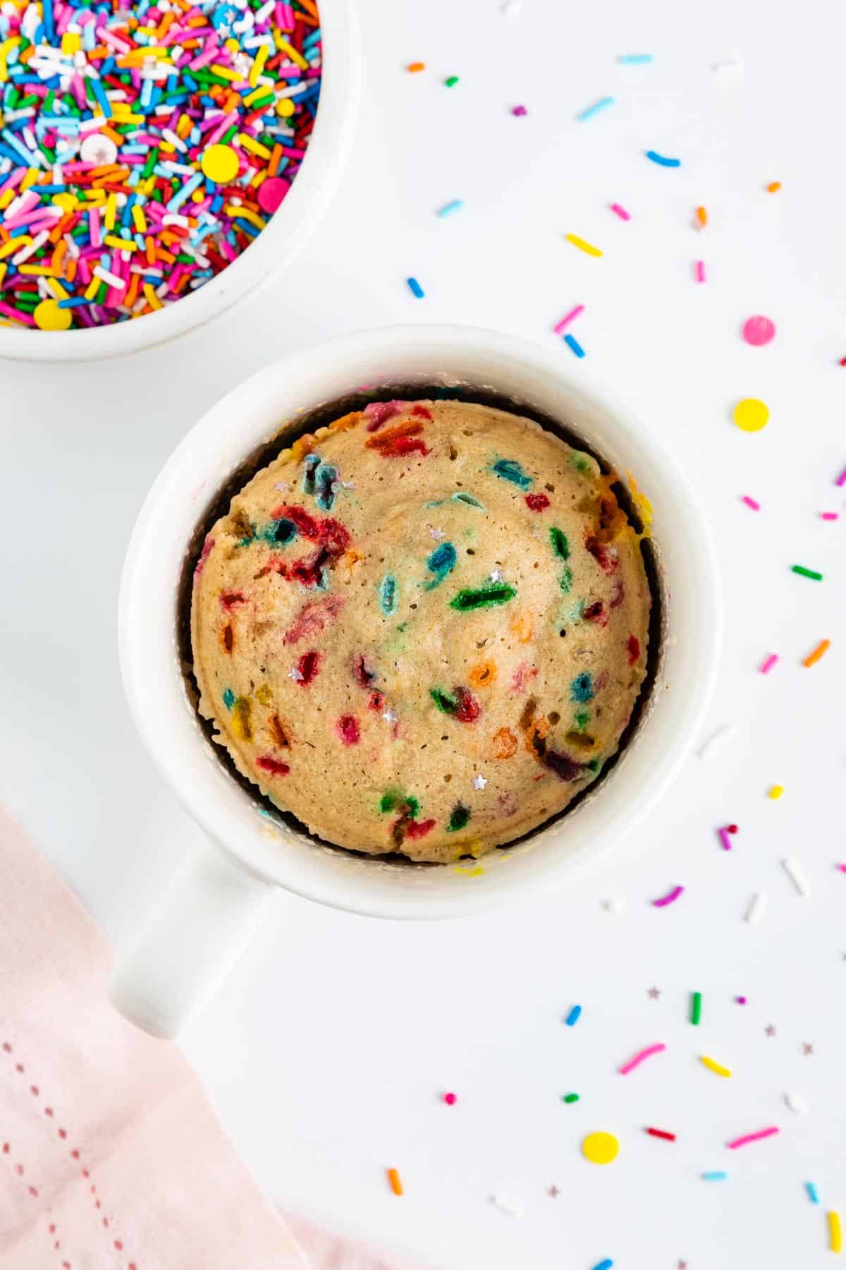 a vegan funfetti mug cake inside a white mug, surrounded by rainbow sprinkles