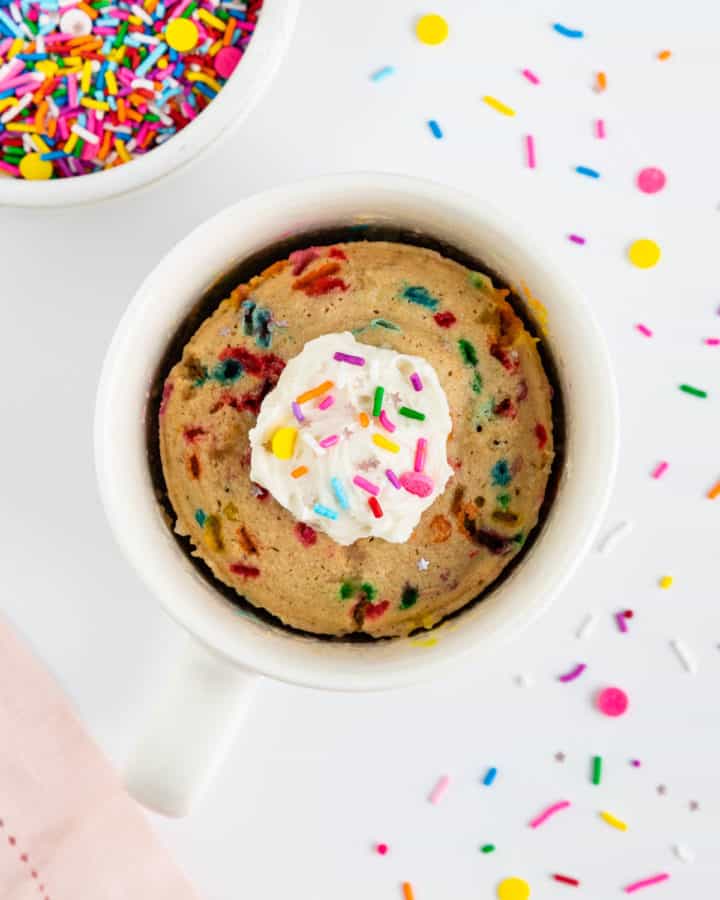 a vegan funfetti mug cake inside a white mug topped with a dollop of vanilla frosting and sprinkles