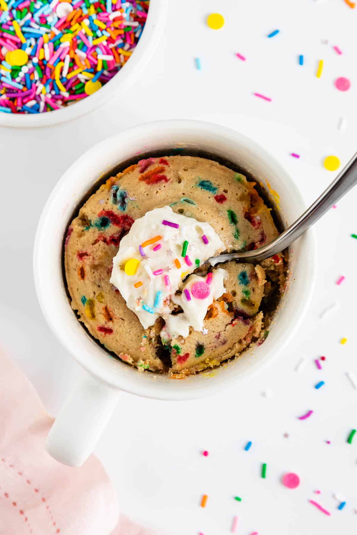 a spoon scooping into a vegan funfetti mug cake with rainbow sprinkles and vanilla frosting