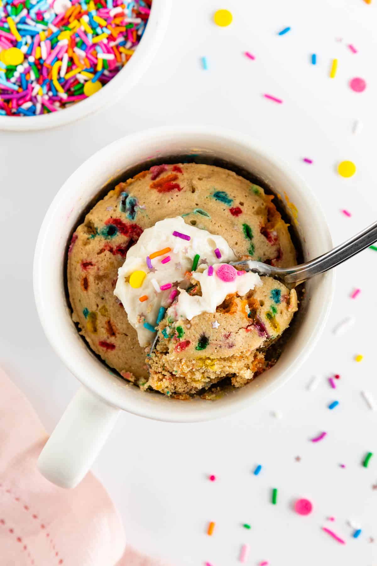 a spoon scooping into a vegan funfetti mug cake with rainbow sprinkles and vanilla frosting