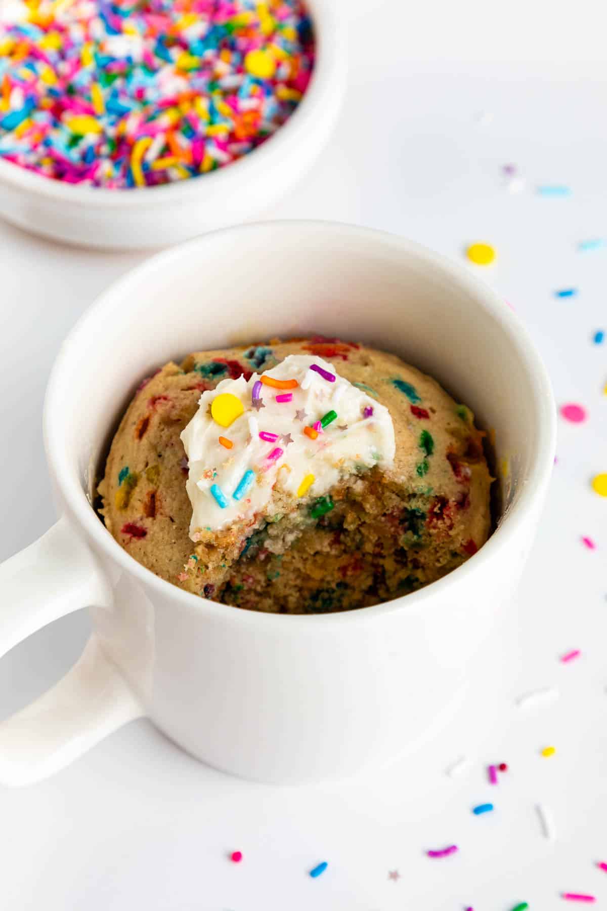 a vegan birthday cake mug cake inside a white mug with vanilla frosting and rainbow sprinkles