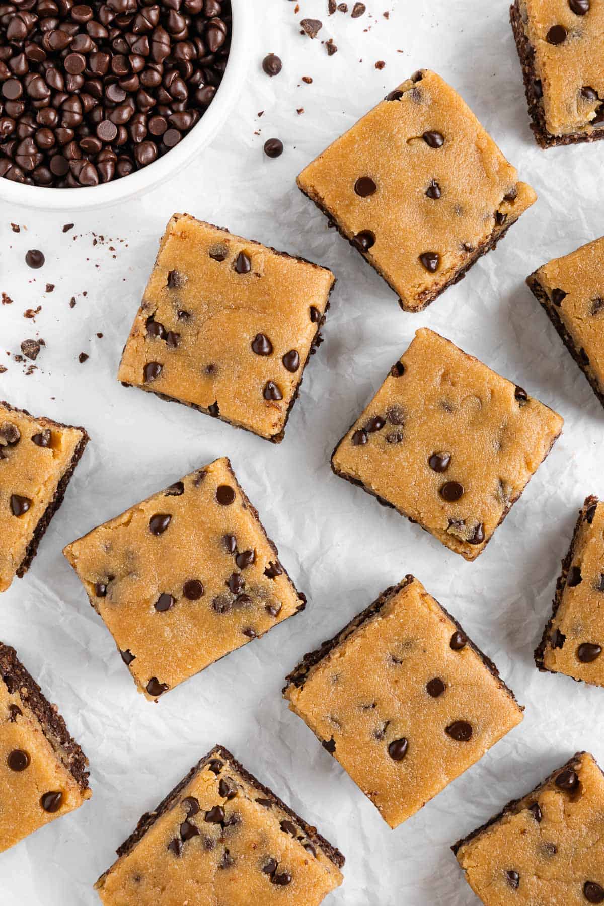 a flat-lay image of no bake cookie dough brownies on white parchment paper beside a bowl of chocolate chips
