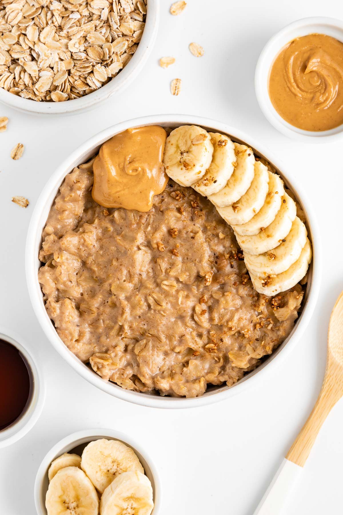 peanut butter oatmeal in a white bowl surrounded by a bowl of maple syrup, a bowl of oats, a bowl of nut butter, and a ceramic mug filled with almond milk