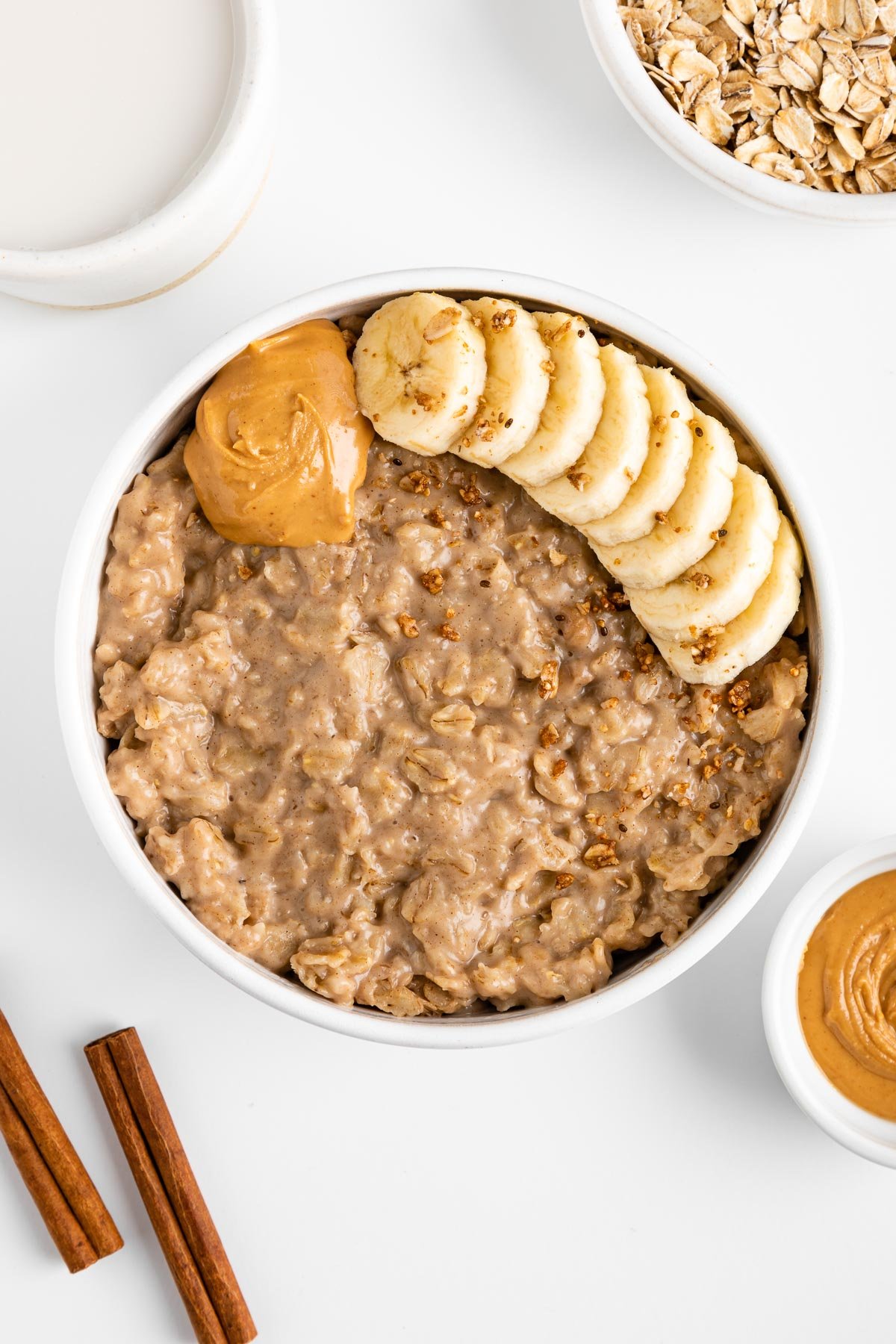 peanut butter oatmeal in a white bowl surrounded by cinnamon sticks, a bowl of oats, a bowl of nut butter, and a ceramic mug filled with almond milk