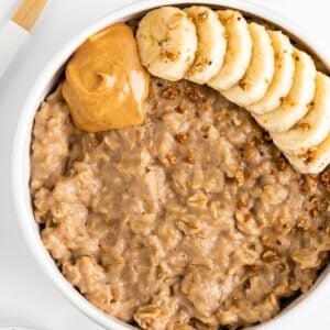 peanut butter oatmeal in a white bowl, topped with sliced banana, granola, and nut butter