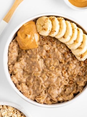 peanut butter oatmeal in a white bowl, topped with sliced banana, granola, and nut butter