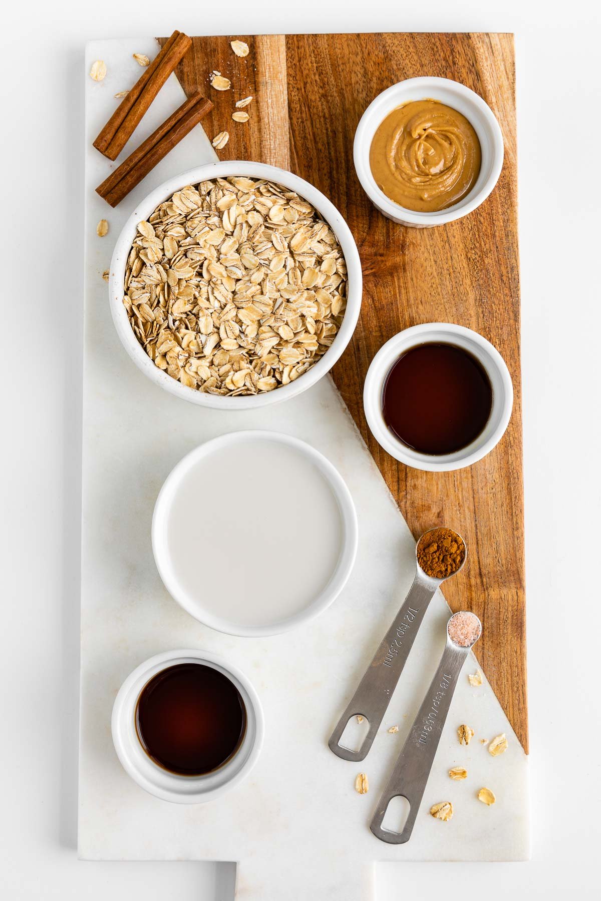 a marble and wood cutting board topped with small white bowls filled with rolled oats, peanut butter, almond milk, maple syrup, vanilla extract, and cinnamon