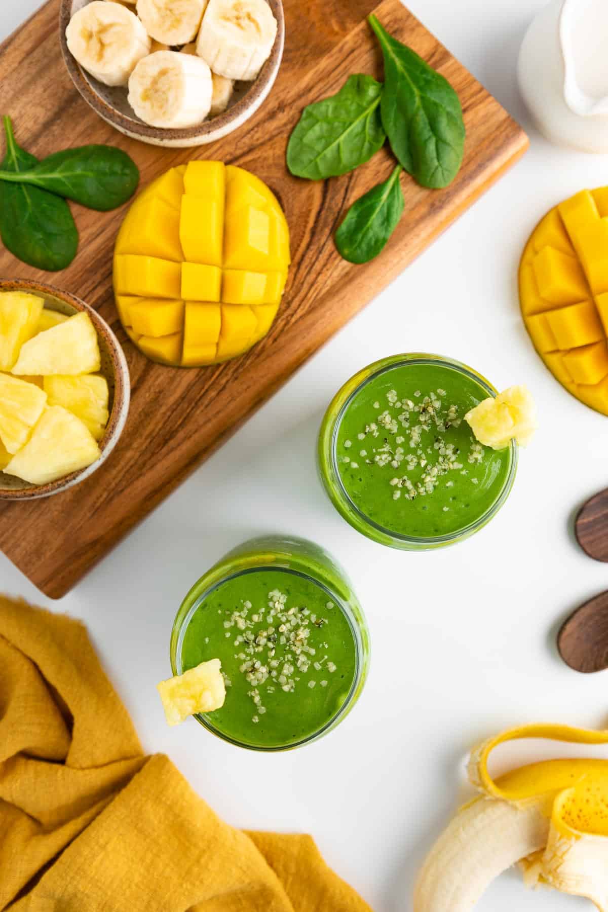 an overhead photo of two green smoothie glasses surrounded by a yellow linen napkin, a peeled banana, mango, a bowl of pineapple, and spinach leaves