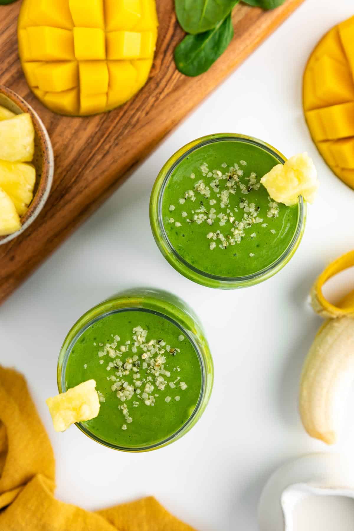 an overhead photo of two green smoothies in glasses, surrounded by sliced mango, pineapple chunks, a peeled banana, spinach, and a yellow linen napkin