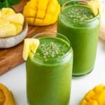 two glasses filled with a green smoothie surrounded by fresh mangoes, banana, pineapple chunks, and spinach