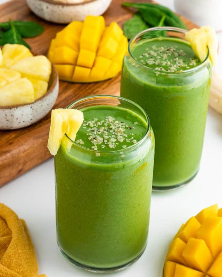 two glasses filled with a green smoothie surrounded by fresh mangoes, banana, pineapple chunks, and spinach
