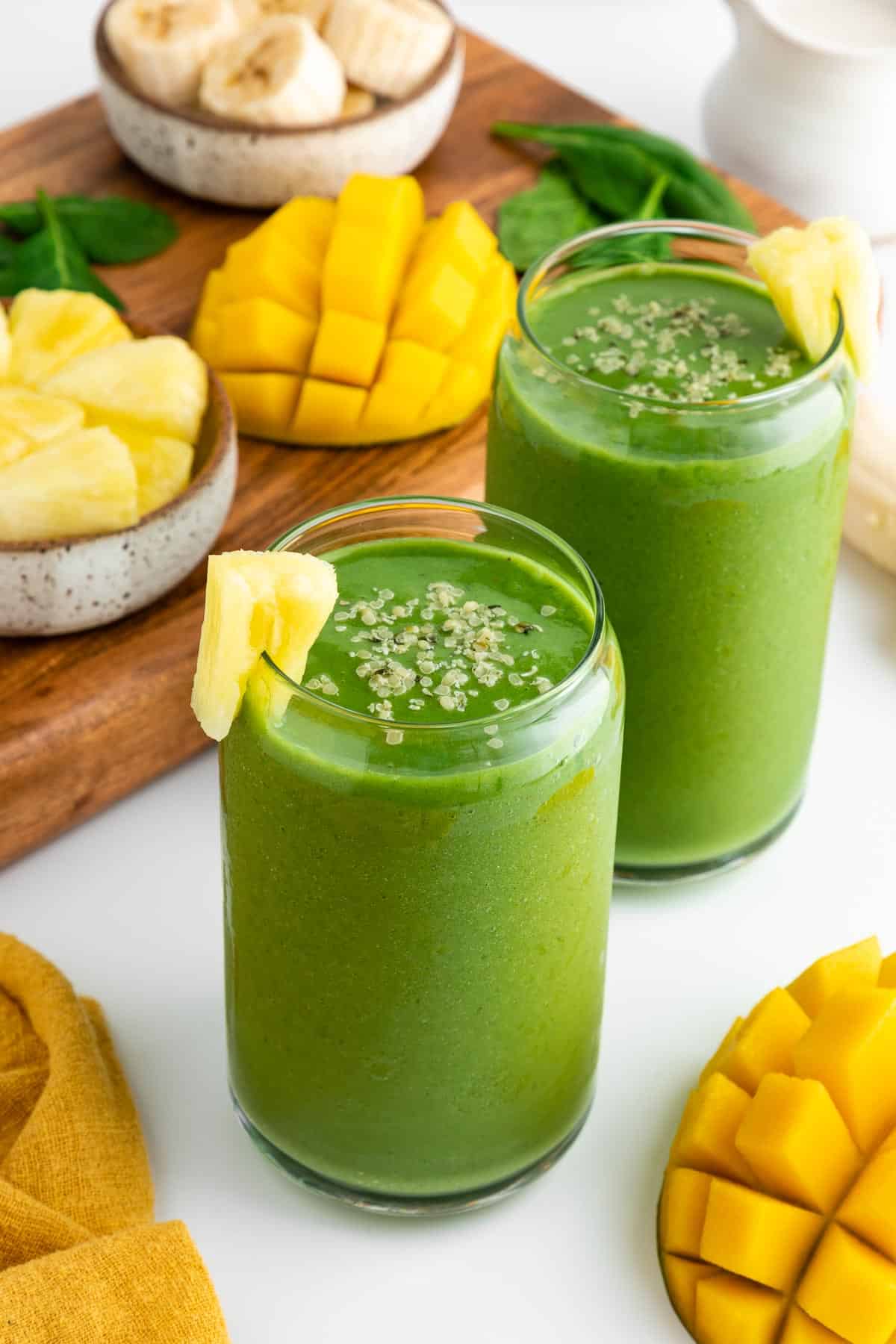 two glasses filled with a green smoothie surrounded by fresh mangoes, banana, pineapple chunks, and spinach