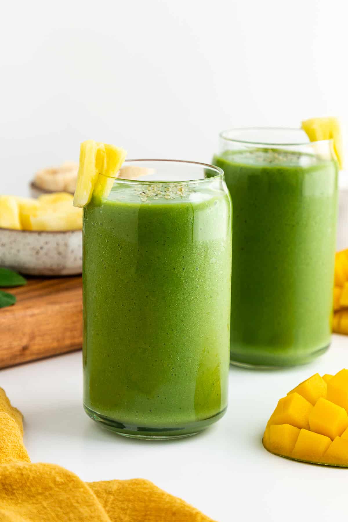 two green smoothies inside glasses beside a bowl of pineapple, two mangoes, and a yellow linen napkin