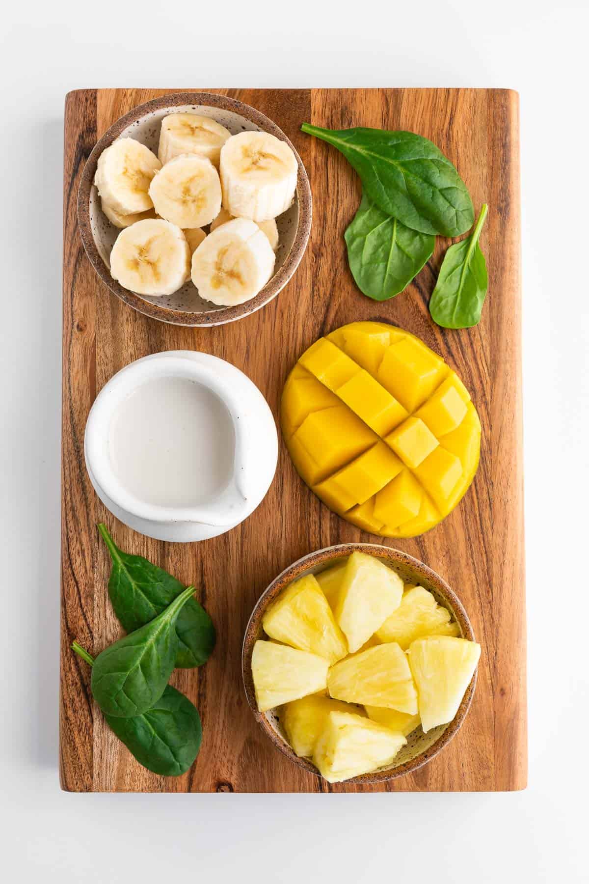 a dark wood rectangular board with a sliced mango, bowl of chopped banana, bowl of pineapple chunks, glass of almond milk, and fresh spinach on top